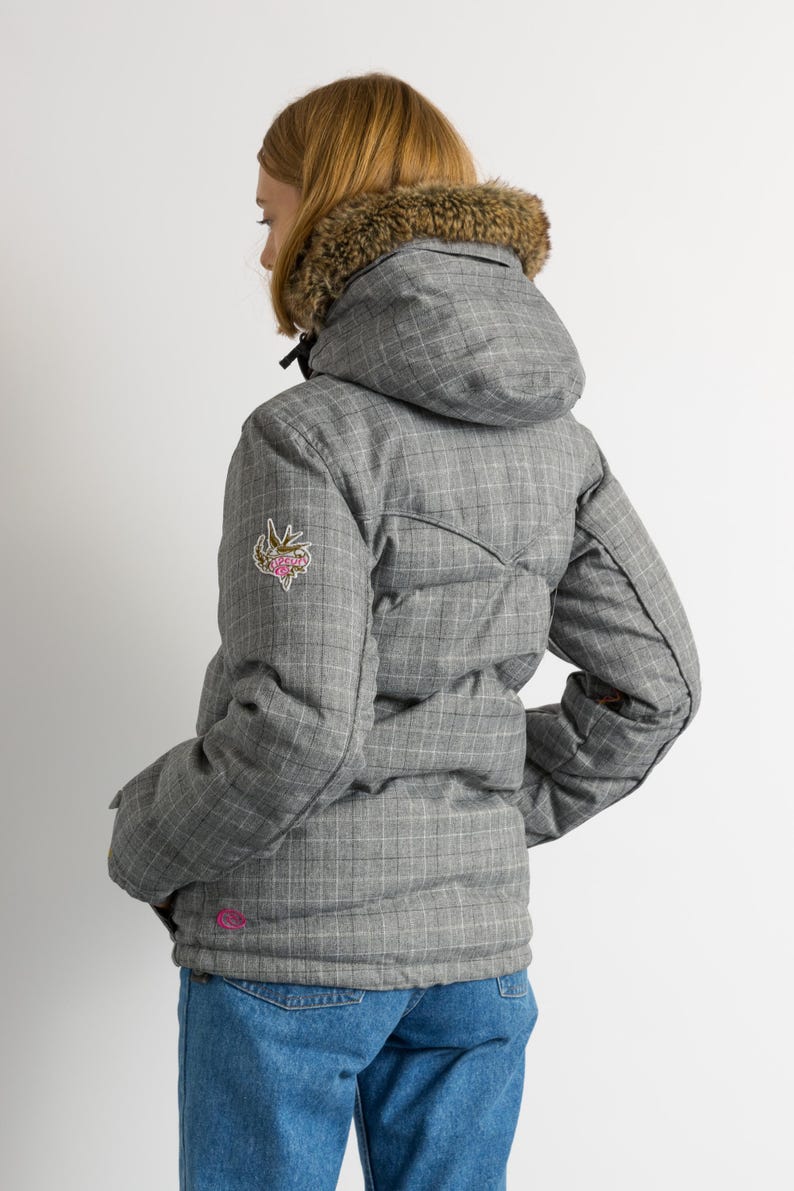 Puede incluir: Chaqueta de invierno acolchada gris con estampado de cuadros y capucha con forro de piel. La chaqueta tiene un peque&ntilde;o parche floral en la manga izquierda y un logotipo rosa cerca del dobladillo. La persona lleva vaqueros azules.