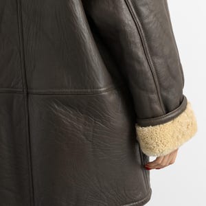 May include: A dark brown leather coat with a shearling lining visible at the cuffs. The coat has a classic design with visible stitching and a textured leather surface. The cuffs are a light tan color.