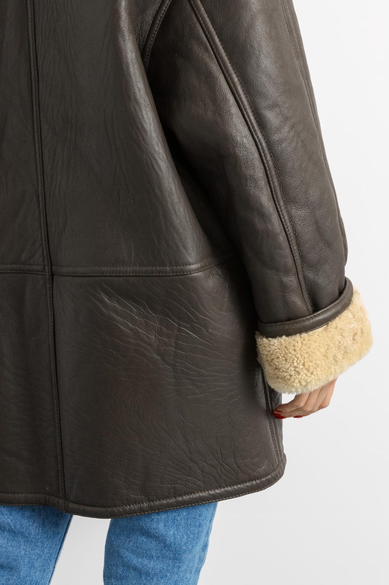 May include: A dark brown leather coat with a shearling lining visible at the cuffs. The coat has a classic design with visible stitching and a textured leather surface. The cuffs are a light tan color.