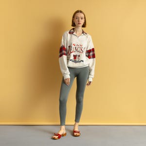 May include: A woman wearing a white sweatshirt with red and black plaid accents and the text "SAN FRANCISCO" on the front. She is wearing gray leggings and red sandals.