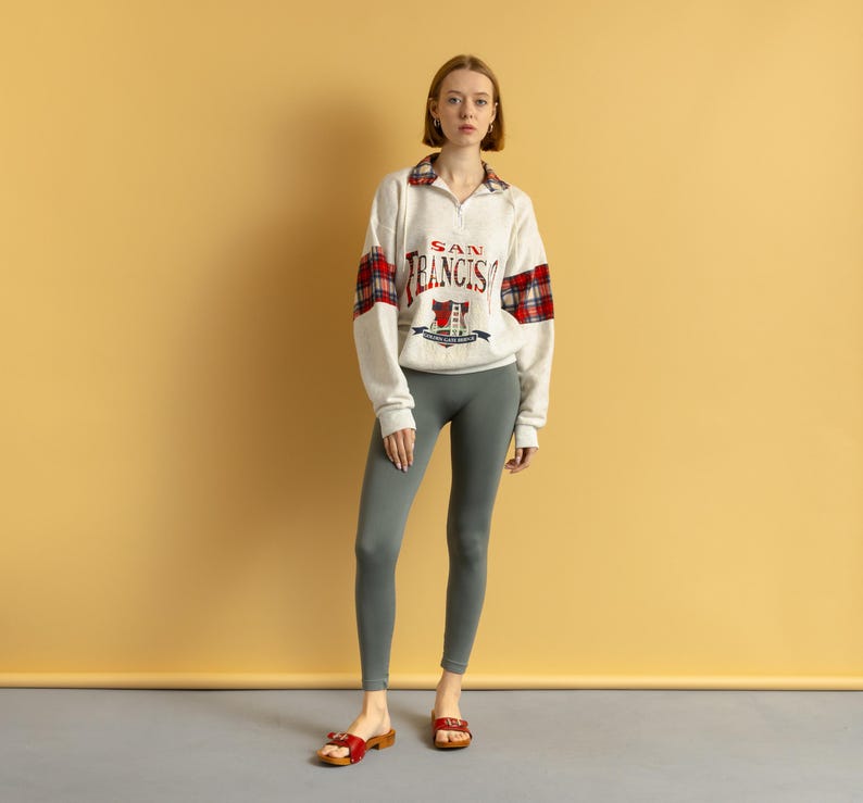 May include: A woman wearing a white sweatshirt with red and black plaid accents and the text "SAN FRANCISCO" on the front. She is wearing gray leggings and red sandals.