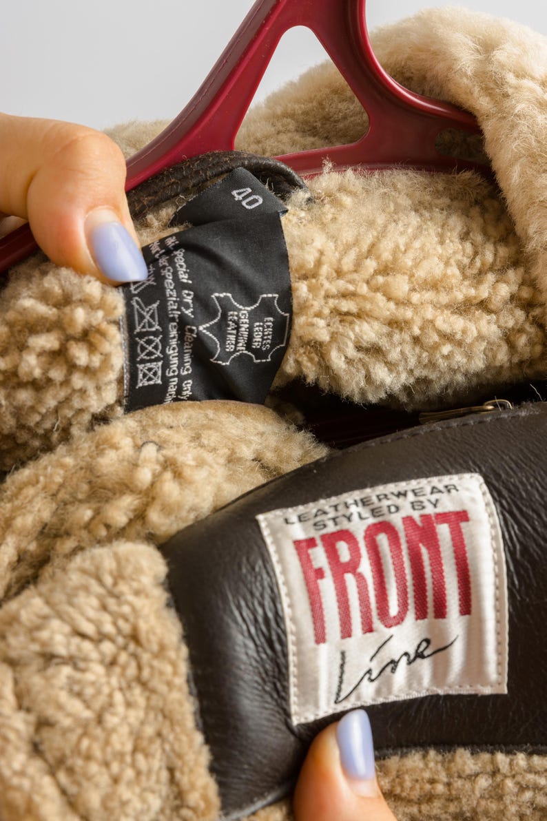 May include: Close-up of a brown leather jacket with a beige shearling lining. The jacket has a label that reads "LEATHERWEAR STYLED BY FRONT Line". A tag shows size 40 and "GENUINE LEATHER".