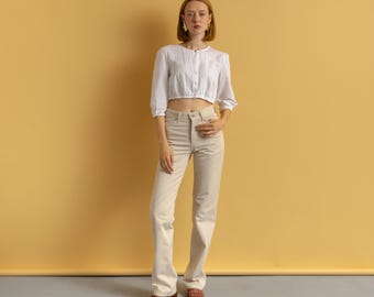 May include: A woman wearing a white, long-sleeved, cropped blouse with pleats and light beige pants. The pants are flared at the bottom. The background is a solid, light orange color.