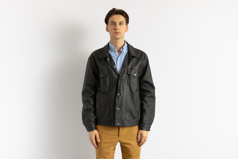 May include: A black leather jacket with a blue button-down shirt underneath. The jacket has a classic design with a collar, button closure, and two pockets.
