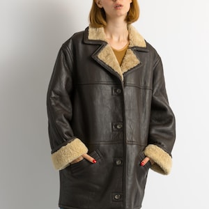 May include: A dark brown leather coat with a beige shearling lining on the collar and cuffs. The coat has a button-up front and two pockets. The model is wearing blue jeans.