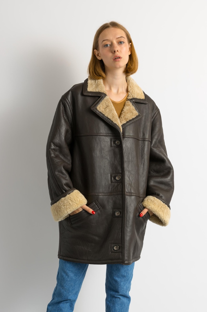 May include: A dark brown leather coat with a beige shearling lining on the collar and cuffs. The coat has a button-up front and two pockets. The model is wearing blue jeans.