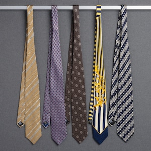 May include: Five neckties of various patterns and colors hang from a silver bar against a gray background. The ties include stripes, polka dots, and floral designs in shades of brown, blue, and purple. Each tie has a label at the bottom.