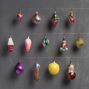 May include: A collection of vintage Christmas ornaments in various shapes and colors, including pink, purple, gold, and green. The ornaments are suspended from thin wires against a gray background, showcasing their intricate designs and reflective surfaces.