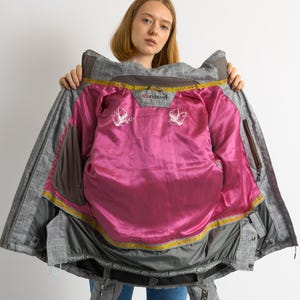 Puede incluir: Chaqueta de invierno a cuadros gris y marr&oacute;n con forro rosa brillante. La chaqueta tiene capucha, m&uacute;ltiples bolsillos y la marca "Rip Curl" es visible. El interior presenta dise&ntilde;os blancos bordados.