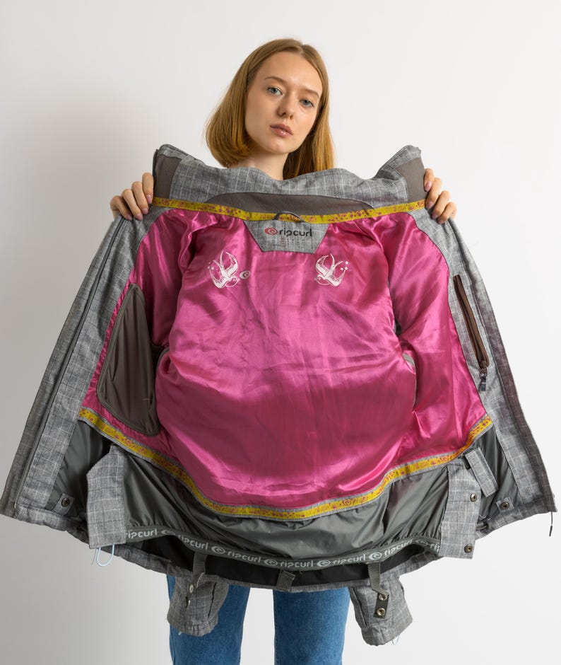 Puede incluir: Chaqueta de invierno a cuadros gris y marr&oacute;n con forro rosa brillante. La chaqueta tiene capucha, m&uacute;ltiples bolsillos y la marca "Rip Curl" es visible. El interior presenta dise&ntilde;os blancos bordados.