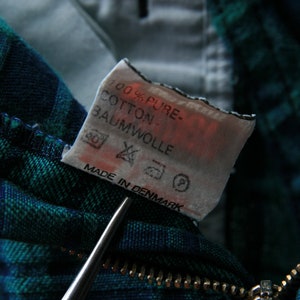 May include: A close-up of a white fabric tag with black text that reads "100% Pure Cotton Baumwolle" and "Made in Denmark". The tag is attached to a green and blue plaid fabric.