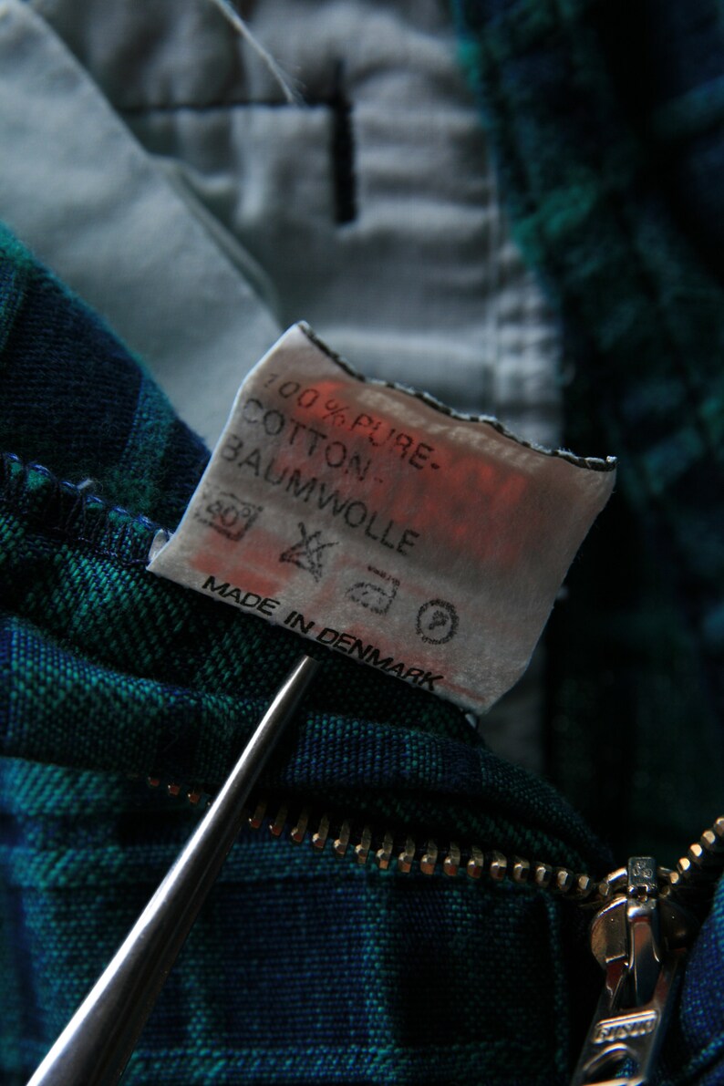 May include: A close-up of a white fabric tag with black text that reads "100% Pure Cotton Baumwolle" and "Made in Denmark". The tag is attached to a green and blue plaid fabric.