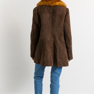 May include: A brown suede coat with a faux fur collar. The coat has a flared hem and is worn with blue jeans and burgundy Chelsea boots. The model is facing away from the camera.