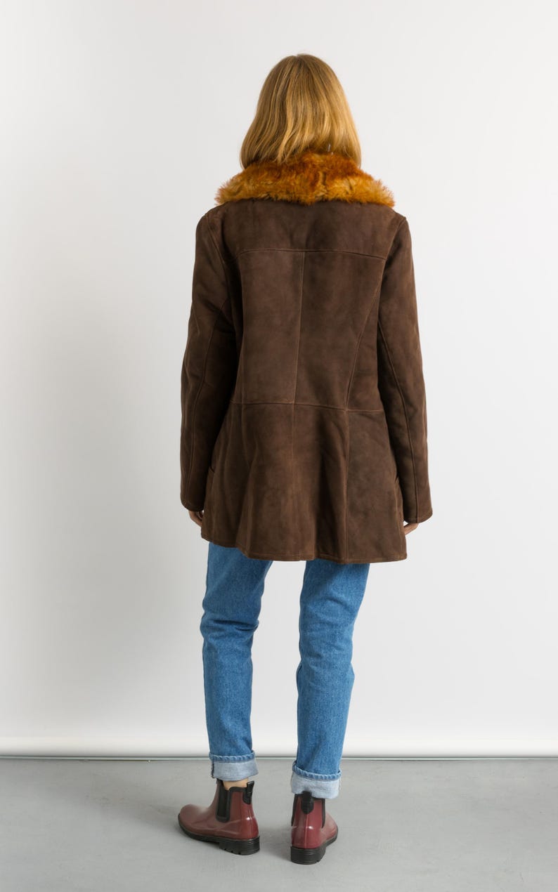 May include: A brown suede coat with a faux fur collar. The coat has a flared hem and is worn with blue jeans and burgundy Chelsea boots. The model is facing away from the camera.