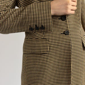 May include: A tan and black houndstooth blazer with black buttons and a pocket. The blazer's pattern is a classic small check design. The sleeve has three buttons.