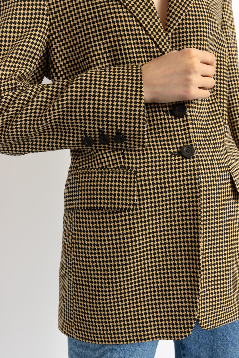 May include: A tan and black houndstooth blazer with black buttons and a pocket. The blazer's pattern is a classic small check design. The sleeve has three buttons.