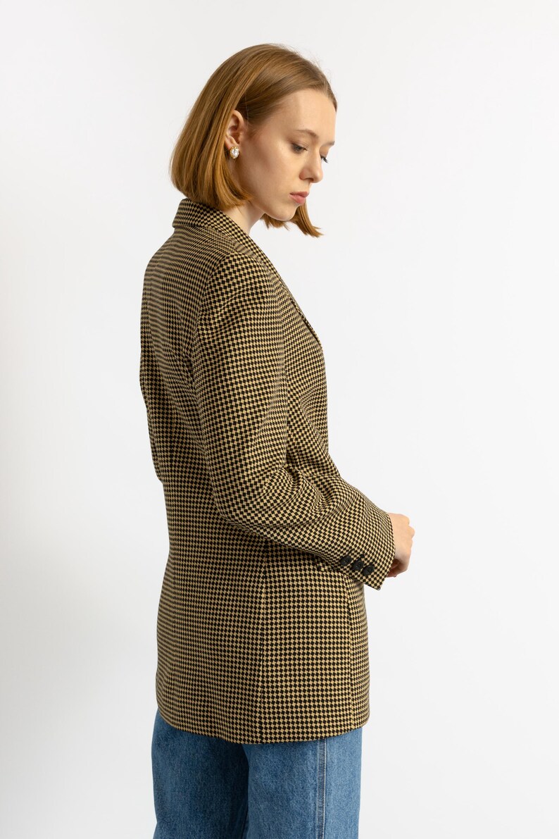 May include: A houndstooth blazer in black and tan. The tailored jacket features a classic pattern, notched lapels, and buttoned cuffs. The blazer is worn with blue jeans.