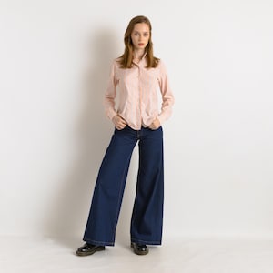 Puede incluir: Una mujer con una camisa de manga larga a rayas rosa y blanca y unos pantalones vaqueros de pierna ancha de color azul oscuro.