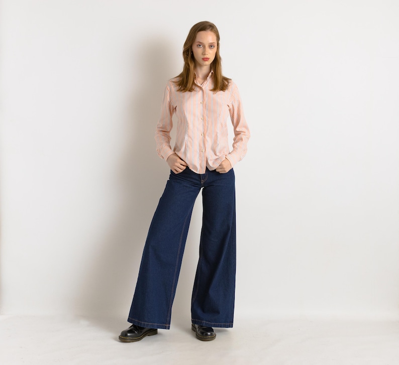 Puede incluir: Una mujer con una camisa de manga larga a rayas rosa y blanca y unos pantalones vaqueros de pierna ancha de color azul oscuro.
