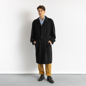 Peut inclure: Un long manteau noir avec une ceinture, porté par une personne aux cheveux bruns. Le manteau a un col et des poches. La personne porte un pantalon marron et des chaussures noires.