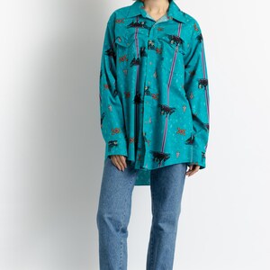 May include: A turquoise blue long-sleeved shirt with a colorful print of abstract shapes and figures. The shirt has a button-down collar and a relaxed fit. The model is wearing the shirt with blue jeans and red clogs.
