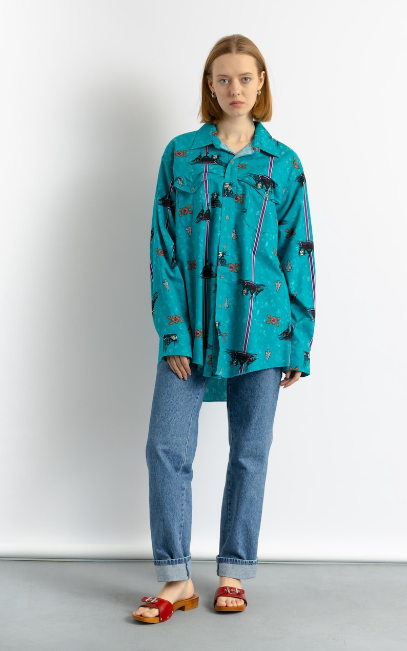 May include: A turquoise blue long-sleeved shirt with a colorful print of abstract shapes and figures. The shirt has a button-down collar and a relaxed fit. The model is wearing the shirt with blue jeans and red clogs.