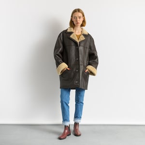 May include: A dark brown shearling coat with a button-up front, beige shearling lining the collar and cuffs. The coat is worn with blue jeans and maroon boots. The coat appears to be vintage or retro style.