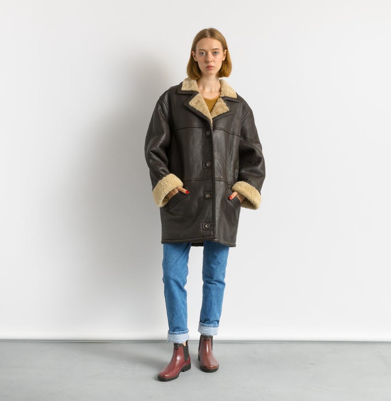 May include: A dark brown shearling coat with a button-up front, beige shearling lining the collar and cuffs. The coat is worn with blue jeans and maroon boots. The coat appears to be vintage or retro style.