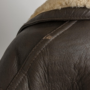 May include: Close-up of a brown leather jacket with a cream-colored shearling collar. The jacket has visible stitching and a textured surface. The image focuses on the shoulder and collar area, showcasing the jacket's design.