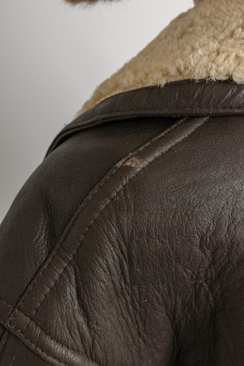 May include: Close-up of a brown leather jacket with a cream-colored shearling collar. The jacket has visible stitching and a textured surface. The image focuses on the shoulder and collar area, showcasing the jacket's design.