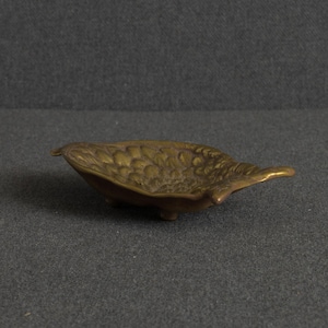 May include: A small, oval-shaped brass dish featuring a textured, leaf-like pattern on its interior surface. The metallic bowl has a warm, aged patina and is supported by three small, integrated feet, giving it a slightly elevated, shallow profile.