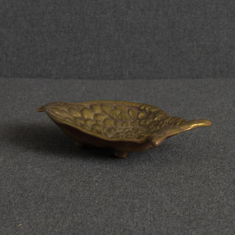 May include: A small, oval-shaped brass dish featuring a textured, leaf-like pattern on its interior surface. The metallic bowl has a warm, aged patina and is supported by three small, integrated feet, giving it a slightly elevated, shallow profile.