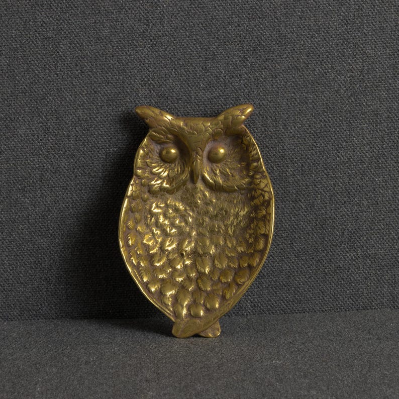 May include: A brass owl-shaped trinket dish featuring detailed embossed feather textures across the body, prominent tufted ears, and two smooth circular eyes. The metal surface displays a warm golden tone with subtle variations in light and shadow.