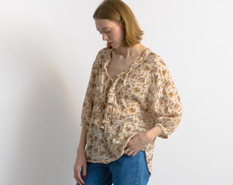 May include: A cream-colored blouse with a floral pattern in shades of brown, purple, and blue. The blouse features a ruffled neckline, three-quarter sleeves, and a tie-up detail at the chest. The model is wearing blue jeans.