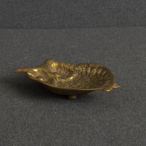 May include: A small, shallow, gold-toned brass dish shaped like an owl. The metal surface features detailed, textured relief patterns representing feathers and facial features, with a smooth, concave interior and small feet on the underside.