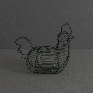 Op de afbeelding: Een zwarte draadmand in de vorm van een kip. De structuur is gevormd door dunne metalen draadspiralen die een hol lichaam creëren met een gedefinieerde kop, snavel en staart. Het ontwerp heeft een klein cirkelvormig oogdetail en een open bovenkant.