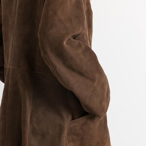 May include: A brown suede coat with a shearling collar. The coat features a classic design with a button closure and pockets. The suede has a soft, matte finish. The shearling collar is a light brown color.