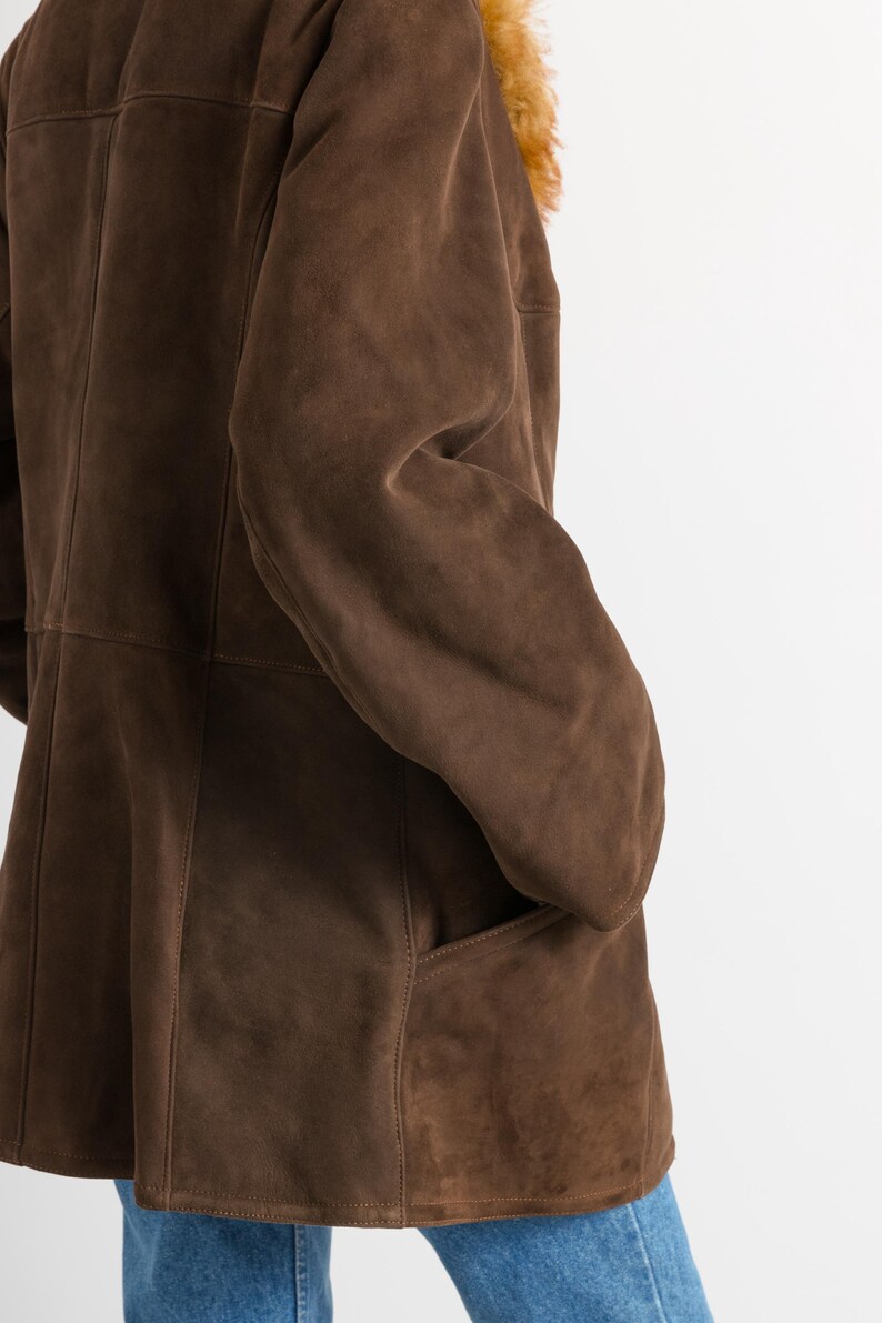 May include: A brown suede coat with a shearling collar. The coat features a classic design with a button closure and pockets. The suede has a soft, matte finish. The shearling collar is a light brown color.
