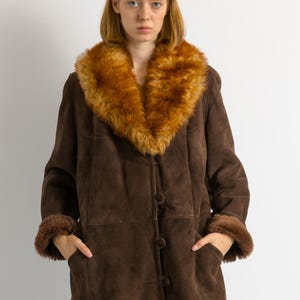 May include: A brown suede coat with a large, fluffy, tan-colored fur collar and cuffs. The coat has a button-up front and pockets. The model is wearing blue jeans.