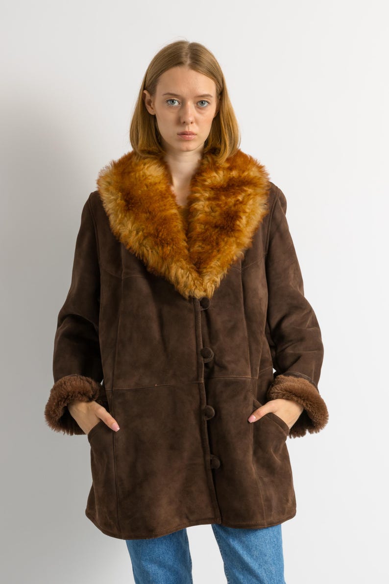 May include: A brown suede coat with a large, fluffy, tan-colored fur collar and cuffs. The coat has a button-up front and pockets. The model is wearing blue jeans.