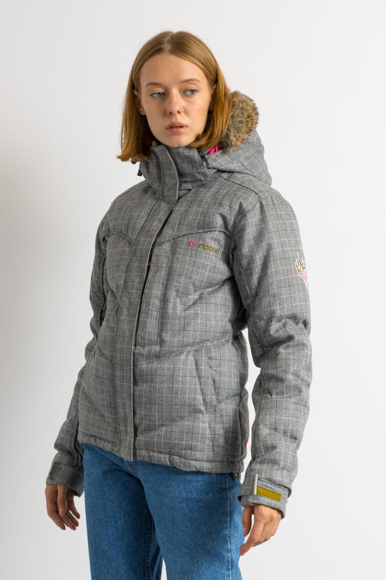 Puede incluir: Chaqueta de invierno gris a cuadros con capucha ribeteada de piel sint&eacute;tica. La chaqueta tiene un forro rosa y el nombre de la marca "Ripcurl" en rosa. Tiene cierre de cremallera y bolsillos. La chaqueta se lleva con vaqueros azules.