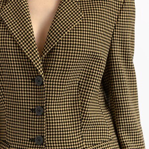 May include: A close-up of a blazer with a black and tan checkered pattern. The blazer has a notched collar, three black buttons, and two front pockets. The fabric has a woven texture.