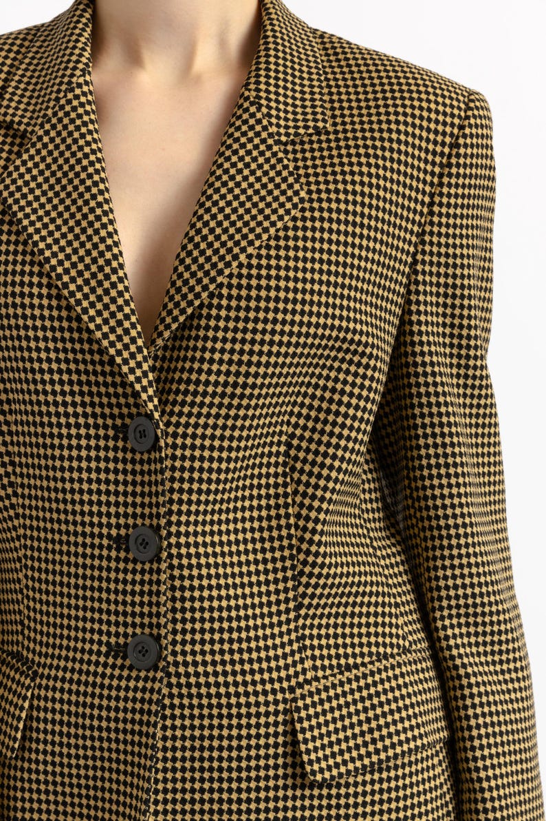 May include: A close-up of a blazer with a black and tan checkered pattern. The blazer has a notched collar, three black buttons, and two front pockets. The fabric has a woven texture.