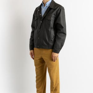 May include: A black leather jacket with a button-up front and a light blue collared shirt underneath. The person is wearing mustard yellow pants and black shoes.