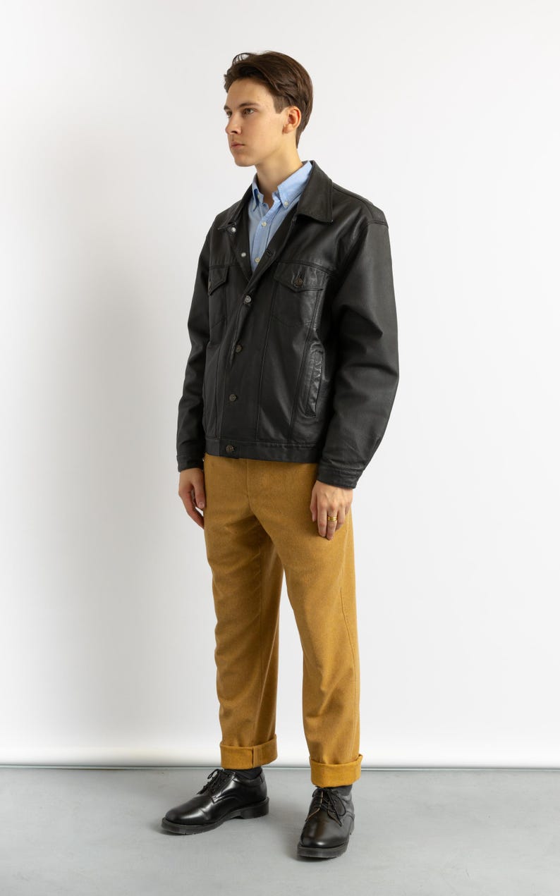 May include: A black leather jacket with a button-up front and a light blue collared shirt underneath. The person is wearing mustard yellow pants and black shoes.