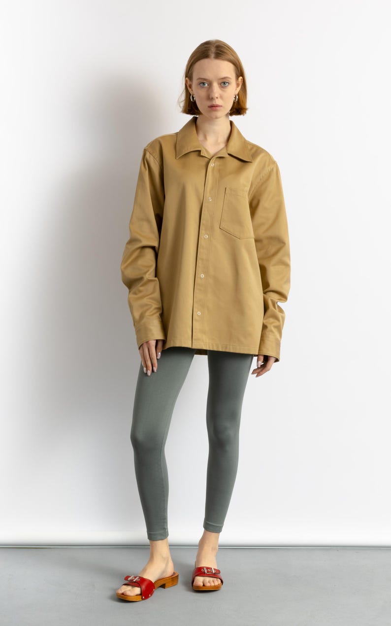 May include: A woman wearing a khaki button-down shirt with a chest pocket, gray leggings, and red wooden clogs.