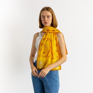 May include: A yellow silk scarf with a brown border and a large brown letter design. The scarf is draped over the shoulders. The scarf has a floral pattern and is worn with a white tank top and blue jeans.