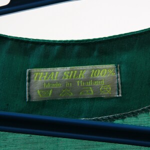 Puede incluir: Una etiqueta de tela de seda verde con el texto "THAI SILK 100%" y "Made in Thailand". La etiqueta tambi&eacute;n incluye instrucciones de cuidado para lavar, secar y planchar.