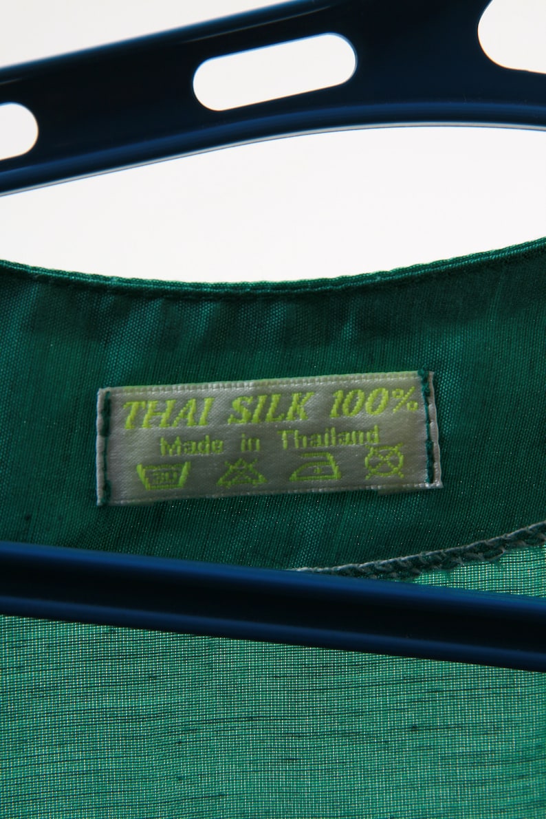 Puede incluir: Una etiqueta de tela de seda verde con el texto "THAI SILK 100%" y "Made in Thailand". La etiqueta tambi&eacute;n incluye instrucciones de cuidado para lavar, secar y planchar.