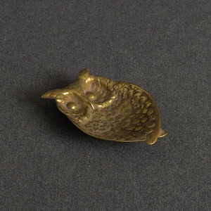 May include: A small, shallow brass dish shaped like an owl. The metal object features a textured surface that mimics feathers, with two prominent circular eyes and pointed ear tufts. The finish is a metallic gold tone with dark patina accents.